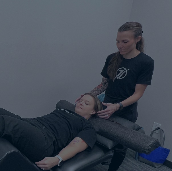 A woman lies on a therapy table with a foam roller under her neck while another woman stands beside her, gently adjusting her head and neck during a treatment session.