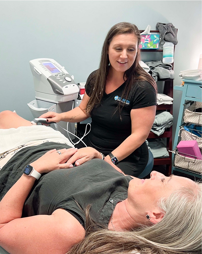 A woman in a black shirt uses a handheld device on another woman lying on a treatment table in a medical or therapy clinic. Both appear relaxed and engaged in conversation. Medical equipment and shelves are visible in the background.