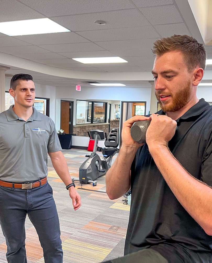 A man in a black shirt holds a dumbbell close to his chest, performing an exercise, while another man in a gray polo shirt observes and instructs him in a gym or physical therapy setting.
