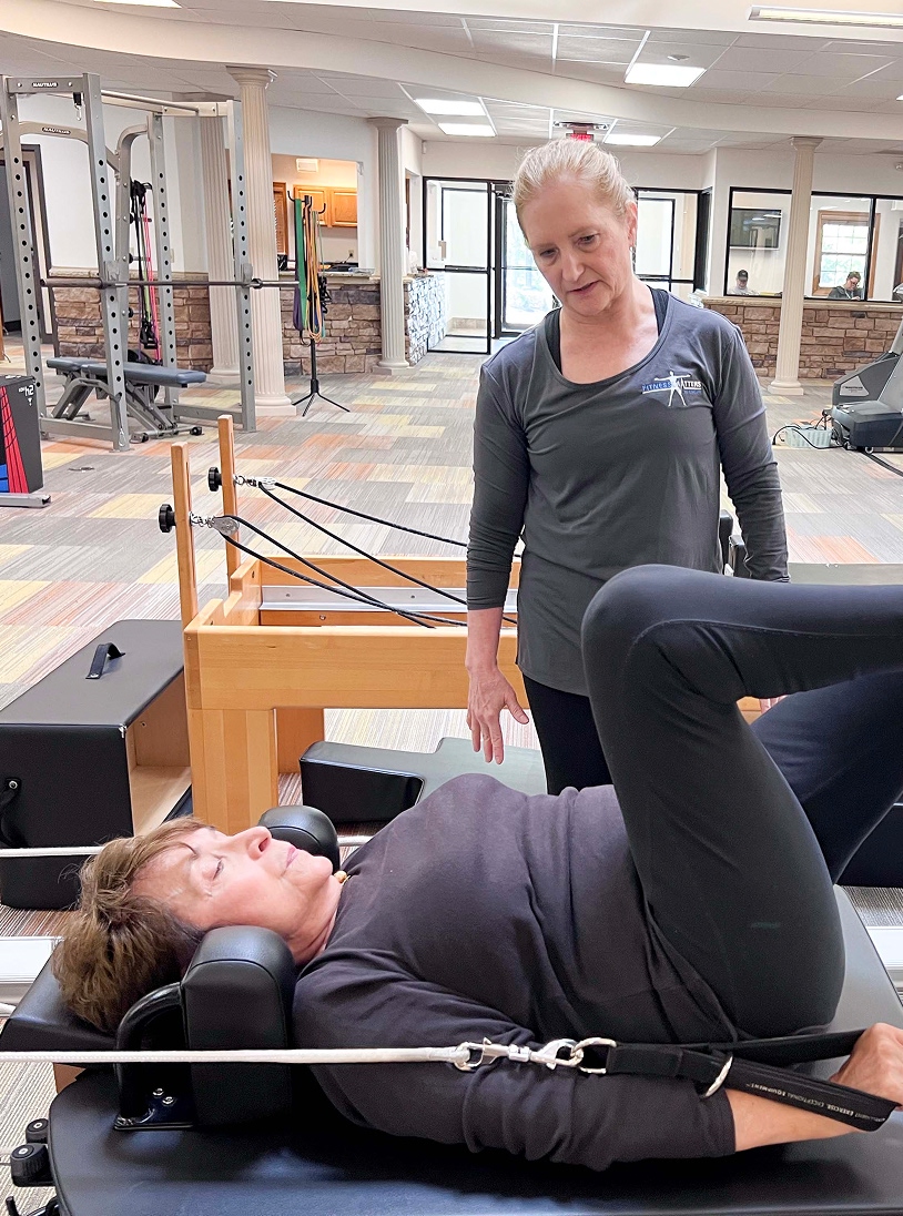 A woman lies on a Pilates reformer, exercising with her knees bent, while an instructor stands beside her, offering guidance in a fitness studio.