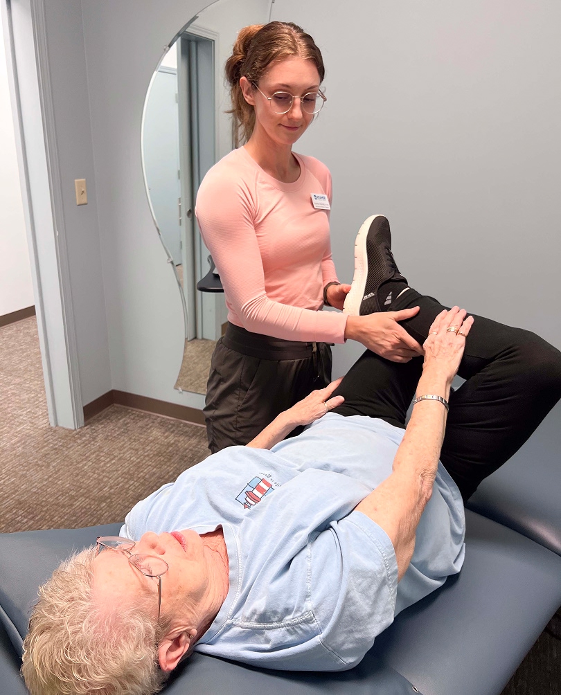 A physical therapist helps an older woman lying on an exam table by gently lifting and bending her leg for a therapy exercise in a clinic room.