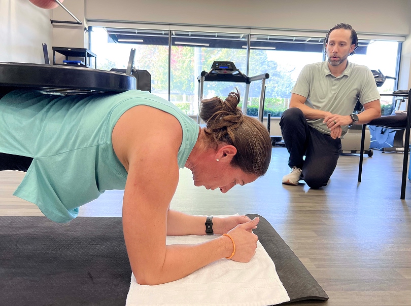 A woman in athletic wear holds a plank position under a weighted platform on her back while a man kneels nearby observing her in a gym with exercise equipment and large windows.