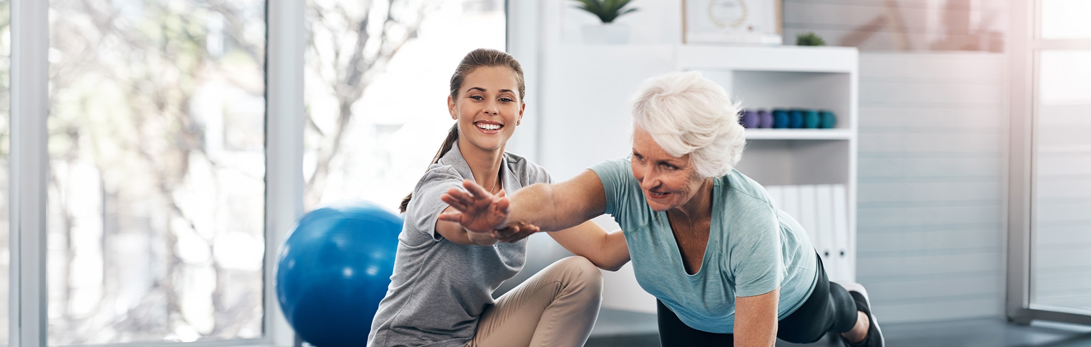 A young woman assists an older woman in a balance exercise indoors; the older woman is stretching forward on one knee while the younger woman supports her, both smiling. A blue exercise ball is in the background.