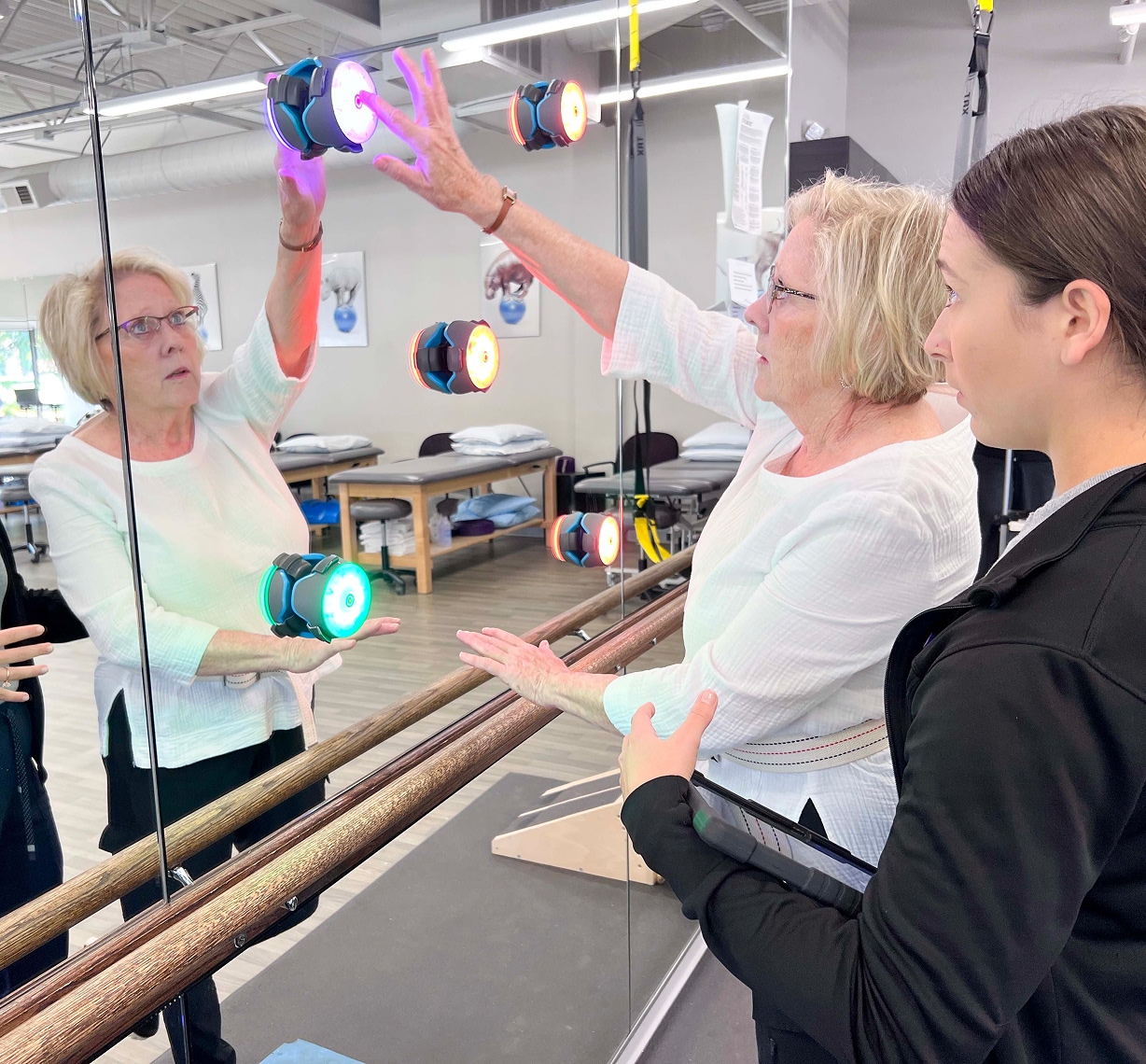 An older woman performs a therapy exercise by touching colored lights on a mirror, while a younger woman observes and takes notes in a rehabilitation clinic.