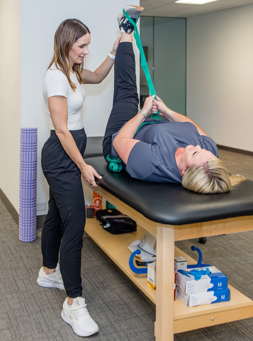 A physical therapist helps a woman lying on a treatment table stretch her leg using a green stretch strap. The therapist supports the woman’s leg while she holds the strap upward. Various medical supplies are under the table.