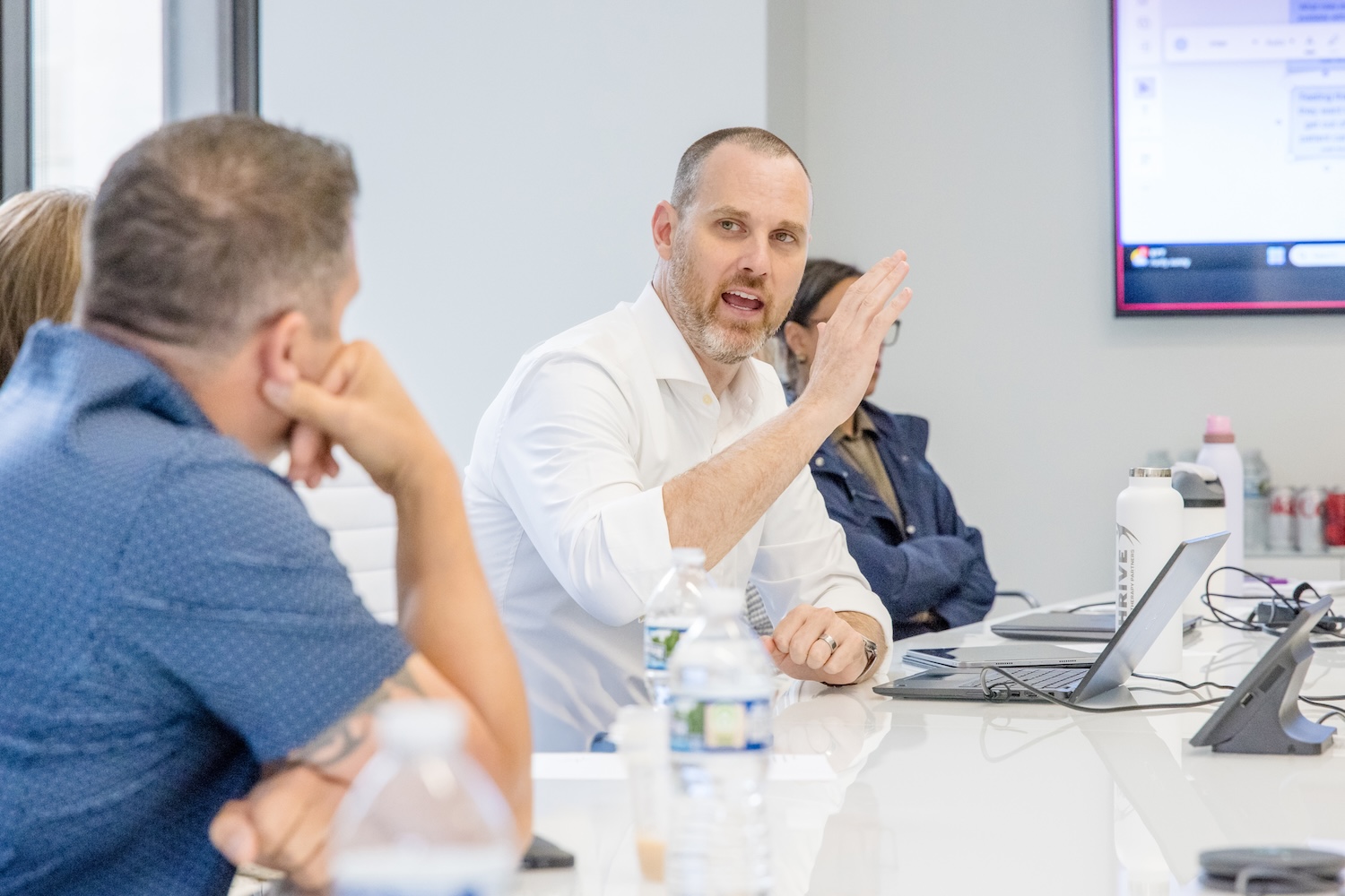 A man in a white shirt gestures while speaking to colleagues during a meeting at a conference table with laptops, water bottles, and papers. A presentation screen is visible in the background.