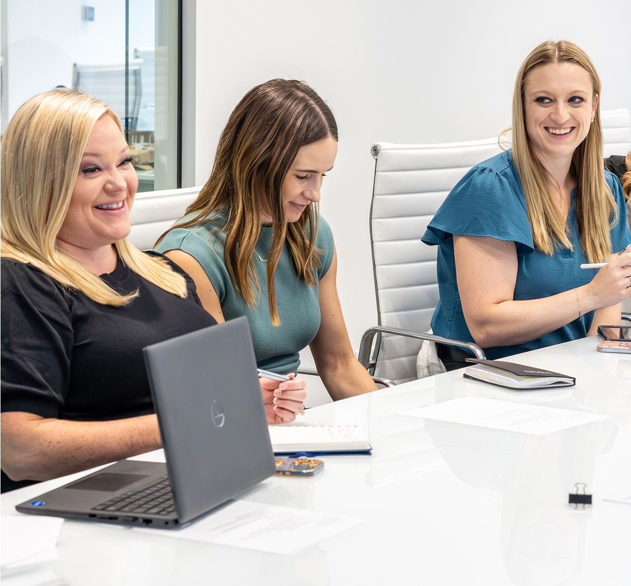 Three women sit at a conference table, smiling and talking. One has a laptop open, and others have notebooks and pens. They appear to be engaged in a positive business meeting in a modern office setting.