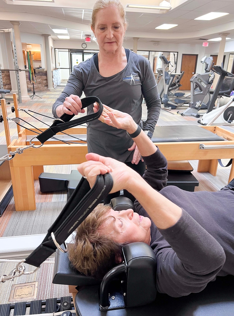 A woman lies on a Pilates reformer machine, holding resistance straps with both hands, while an instructor stands beside her, offering guidance in a bright, modern exercise studio.