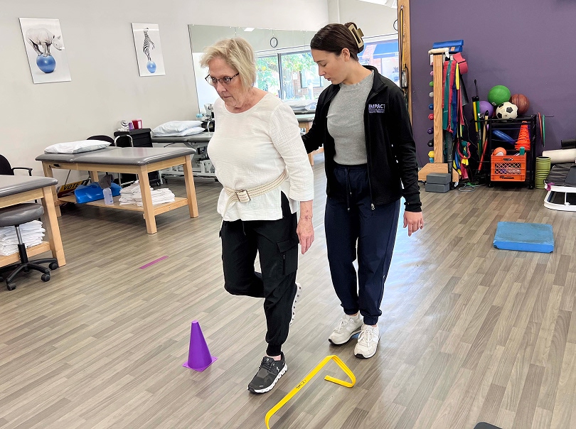 An older woman steps over a yellow hurdle during a physical therapy session, guided by a therapist. Exercise equipment and treatment tables are visible in the background.