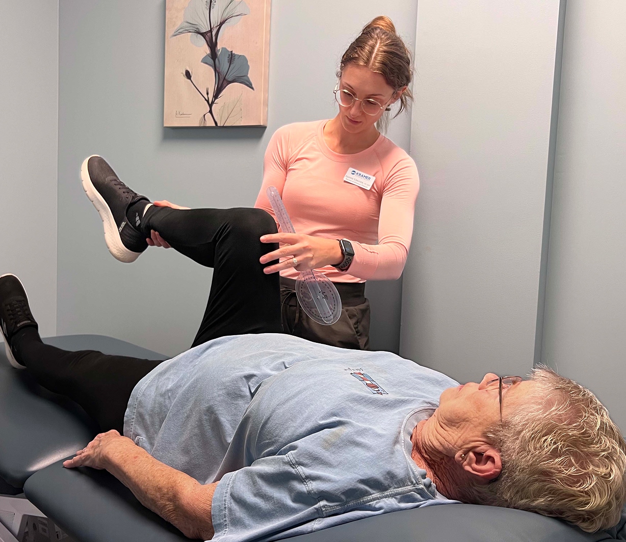 A healthcare professional measures the knee angle of an older adult lying on an exam table using a goniometer. The patient wears glasses and casual clothes; the professional holds the patient's bent leg and records the angle.