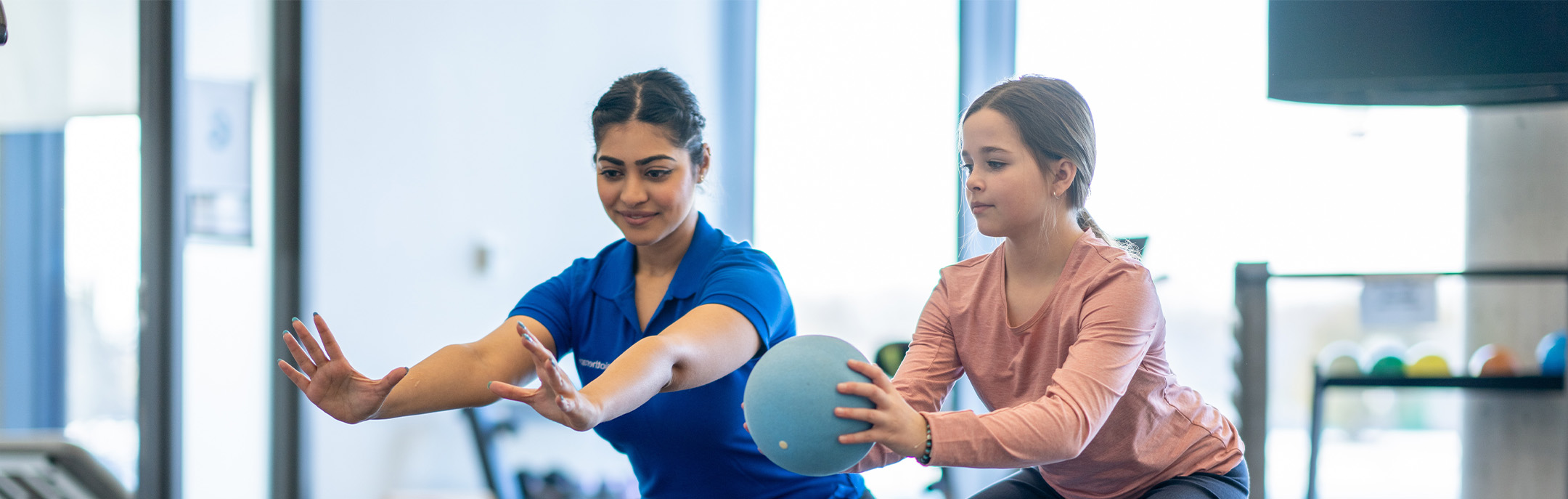 A woman guides a young girl holding a ball during an exercise session in a gym. Both are focused, extending their arms forward, with gym equipment visible in the background.