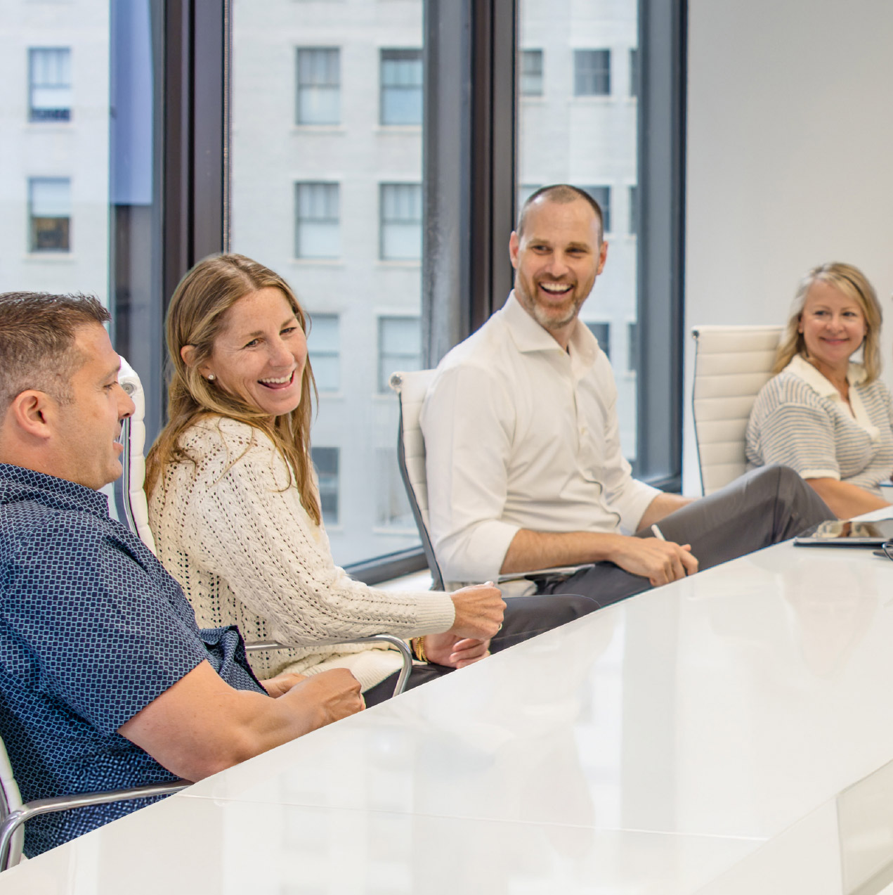 Four people sit around a white conference table, smiling and talking. Large windows behind them show an urban view. The atmosphere appears relaxed and friendly.