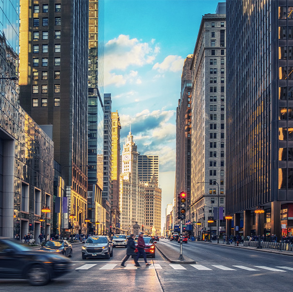 A busy city street scene with tall buildings on both sides, cars driving and parked, and people crossing a wide crosswalk under a partly cloudy, sunny sky. The evening light casts a warm glow on the buildings.