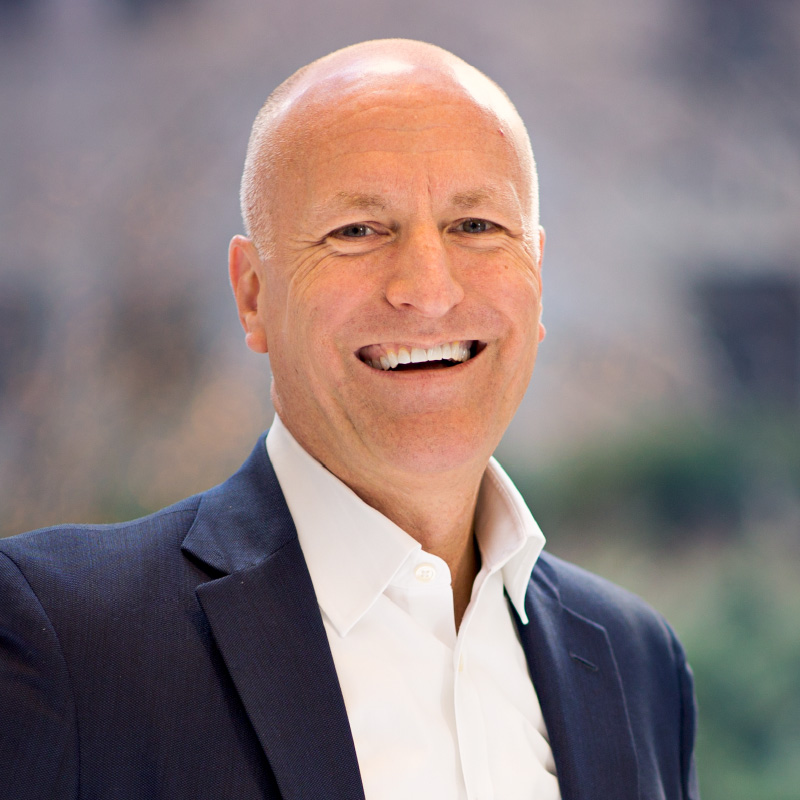 A bald man in a navy suit and white shirt smiles broadly, standing against a softly blurred outdoor background.