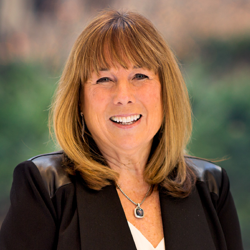 A woman with shoulder-length light brown hair and bangs smiles, wearing a black blazer with leather patches on the shoulders and a silver pendant necklace, standing in front of a blurred green background.