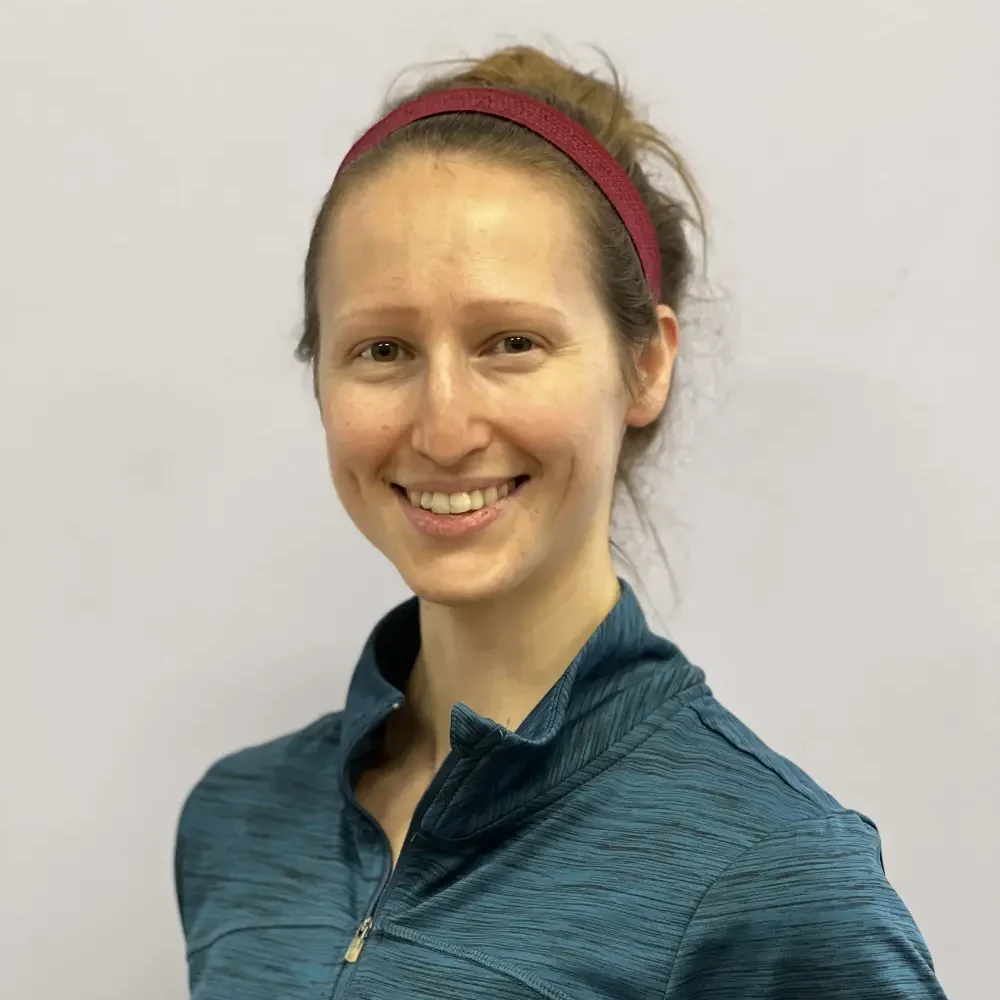 A woman with light skin, wearing a blue zip-up athletic top and a red headband, smiles at the camera against a plain light background.