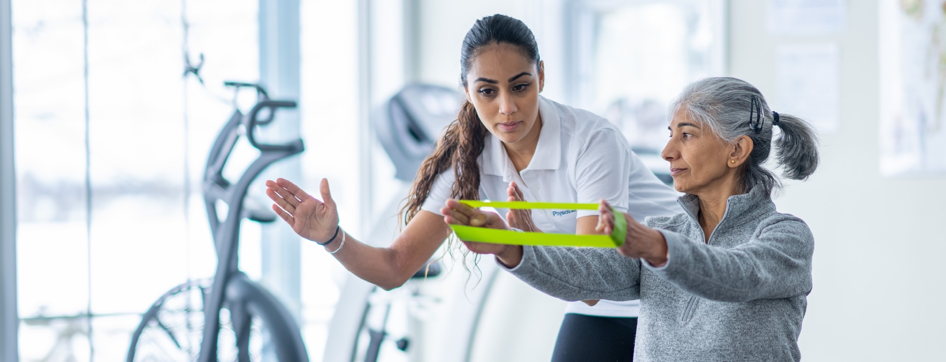A physical therapist assists an older woman using a yellow resistance band in a bright exercise room with fitness equipment in the background.