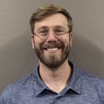 A man with short light brown hair, a beard, and glasses is smiling at the camera. He is wearing a blue collared shirt and is standing in front of a plain gray wall.