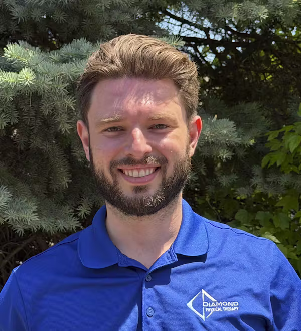 A man with brown hair and a beard is smiling outdoors, wearing a blue "Diamond Physical Therapy" polo shirt. Green trees are in the background.
